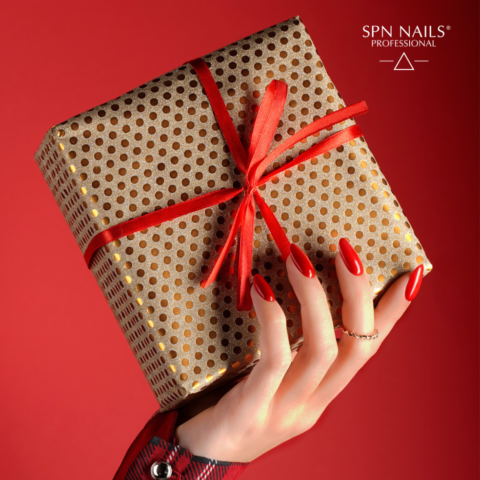 Dłoń trzymająjca zapakowany prezent. Paznokcie pomalowane lakierem hybrydowym w kolorze czerwonym z drobinkami